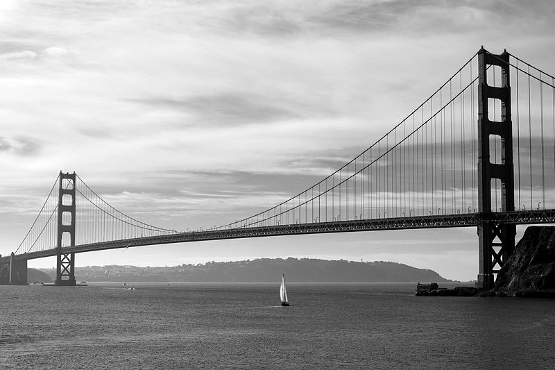 Suspension bridge in black and white with a single sail beneath it