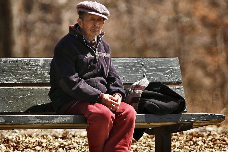 Man in a flat cap sitting alone on a park bench among fallen leaves
