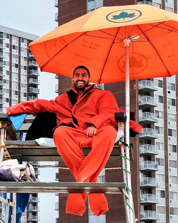 Lifeguard seated beneath an orange umbrella, tower blocks behind